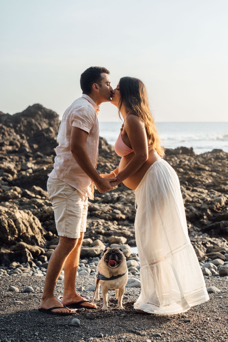 Expecting couple kissing on a pebble beach, with their pug at their feet